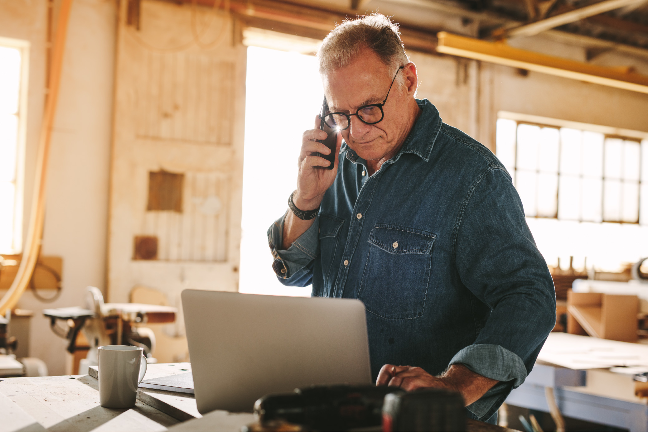 iStock-1135158324_Senior-man-talking-on-cell-phone-and-using-laptop-on-work-table.-Mature-carpenter-working-on-laptop-and-answering-phone-call-in-his-carpentry-workshop_RESIZED